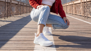 Nahaufnahme der Füße einer Person in roter Jacke und heller Jeans, die in der Hocke auf einer Holzbrücke mit weißer Markierungslinie sitzt. Sie trägt komplett weiße, sportliche Sneaker.