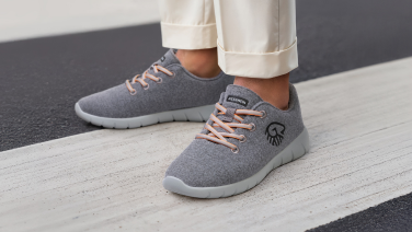 Nahaufnahme der Füße einer Person in cremefarbener, hochgekrempelter Hose. Sie trägt graue Merino Wolle Sneaker mit apricotfarbenen Schnürsenkeln und hellgrauer Sohle. Die Person steht auf einem Zebrastreifen.