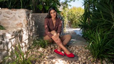 Frau in braunem Anzug sitzt vor Steinmauer in mediterranem Garten, trägt rote Giesswein Ballerinas
