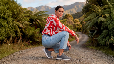 Eine Frau in rot-weiß gemustertem Oberteil und heller Jeans hockt auf einem Schotterweg, umgeben von üppiger grüner Farnvegetation und Bergen im Hintergrund. Sie trägt graue Merino Wolle Sneaker.