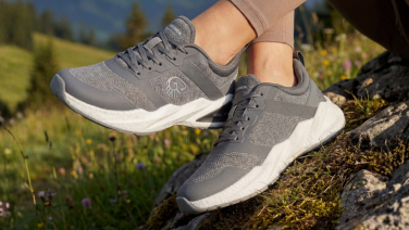 Nahaufnahme der Füße einer Person, die auf einem moosbewachsenen Felsen in einer Berglandschaft steht. Sie trägt graue, sportliche Sneaker mit weißer Laufsohle und sichtbarem Logo an der Zunge.