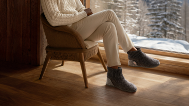 Person in beiger Freizeitkleidung mit dunkelgrauen Giesswein Wollhausschuhen sitzt entspannt am Fenster mit Blick auf verschneite Winterlandschaft