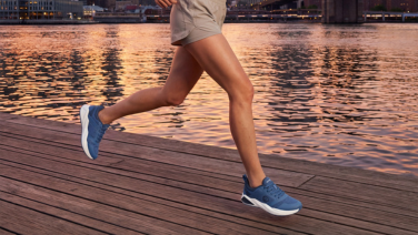 Eine Person in beiger Sportbekleidung läuft über einen Holzsteg am Wasser bei Sonnenuntergang, die Umrisse einer Stadt sind im Hintergrund zu erkennen. Sie trägt blaue, sportliche Laufschuhe.