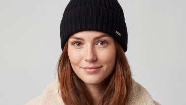 Porträt einer lächelnden Frau mit rotbraunem Haar und schwarzer Rippstrick-Mütze mit Metall-Label, sie trägt eine cremefarbene Teddyfleece-Jacke vor hellgrauem Hintergrund.