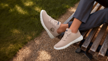 Nahaufnahme beigefarbener Giesswein Merino-Sneaker mit weißer Sohle und Logoprägung an den Füßen einer Person in grauer Hose auf einer Parkbank.