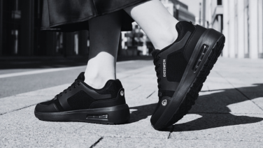 Schwarz-Weiß-Nahaufnahme eines schwarzen Giesswein Sneakers mit Luftkammersohle an den Füßen einer Person auf einem Stadtpflaster.