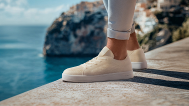 Nahaufnahme cremefarbener Giesswein Merino-Sneaker mit weißer Flachsohle auf einer Steinmauer vor mediterraner Meereskulisse.