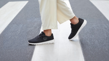 Nahaufnahme schwarzer Giesswein Merino-Sneaker mit weißer Sohle an den Füßen einer Person in weißer Weithose auf einem Zebrastreifen.