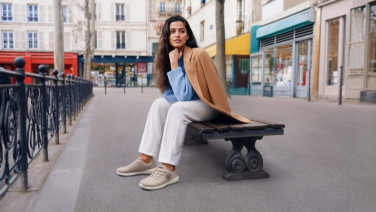 Frau sitzt auf städtischer Bank auf Platz in Innenstadt, trägt beige Jacke, blaues Shirt, weiße Hose und beige Giesswein Merino Runners Sneaker