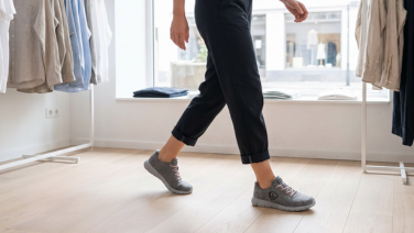 Eine Person in dunkler, hochgekrempelter Hose geht durch einen hellen Laden mit Holzboden, im Hintergrund hängen Kleidungsstücke an Ständern. Sie trägt graue Merino Wolle Sneaker mit Schnürung.