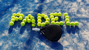 Ein schwarzer Padel-Schläger liegt auf einem blauen Hartplatz. Gelb-grüne Tennisbälle sind daneben zum Schriftzug „PADEL" angeordnet.