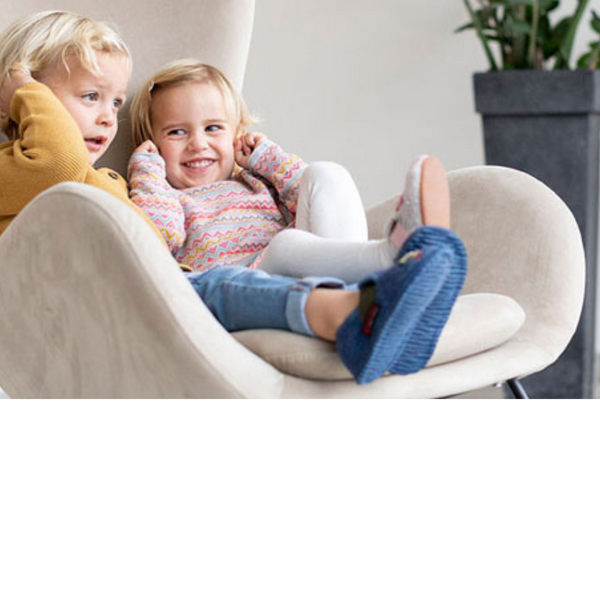 Zwei Kleinkinder sitzen gemeinsam in einem beigen Sessel, das vordere Kind trägt blaue gestrickte Hausschuhe und lächelt fröhlich
