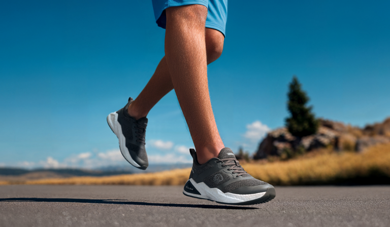 Nahaufnahme von Beinen in grauen Giesswein Laufschuhen mit weißer Sohle in Laufbewegung auf einer Straße. Die Person trägt ein blaue Shorts und läuft vor klarem blauem Himmel. Im Hintergrund verschwommen Berge und ein Baum.