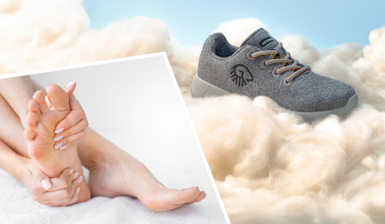 Ein grauer Merino-Sneaker mit hellgrauer Sohle und Schnürsenkeln (Merino Runners von Giesswein) liegt eingebettet in weicher, wolkenartiger Wolle vor blauem Himmel – Symbol für Leichtigkeit und Komfort. Im unteren Bildabschnitt hält eine Person mit gepflegten Händen ihren Fuß schützend in der Hand – Hinweis auf Fußgesundheit und Wohlbefinden.