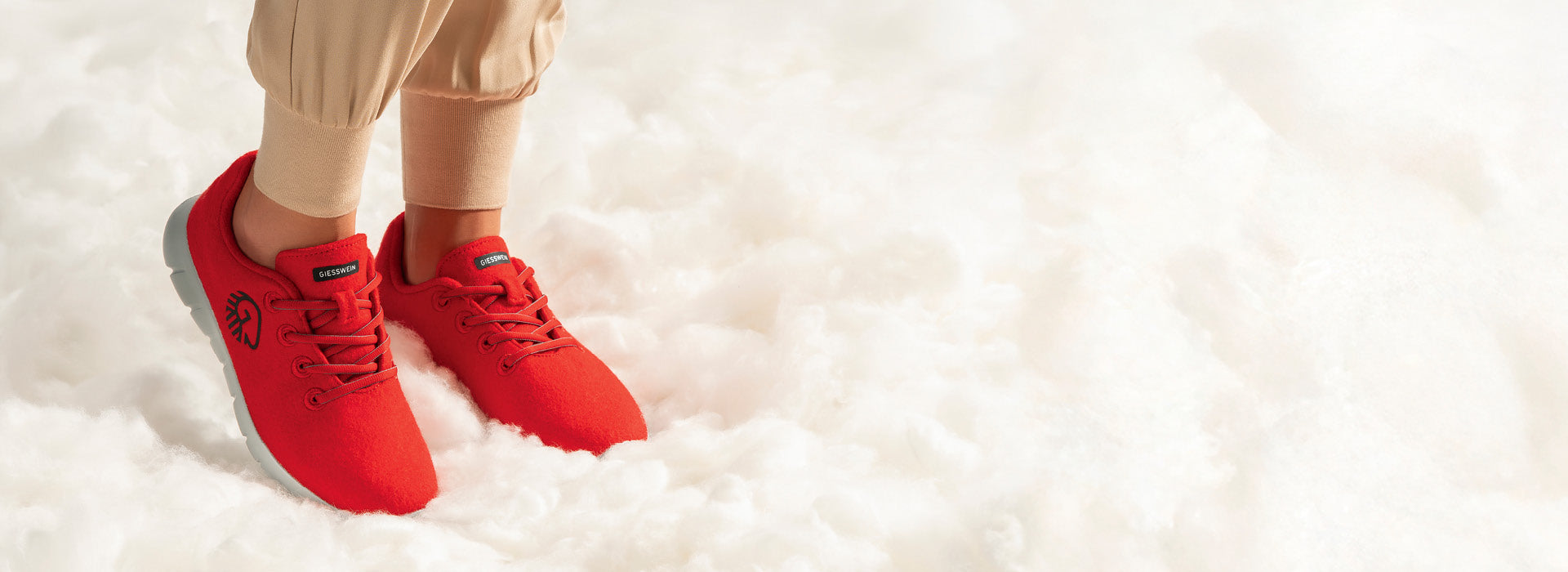 Eine Person trägt leuchtend rote Giesswein-Sneaker mit grauer Sohle (Merino Runners von Giesswein). Die Schuhe bestehen aus weichem Stoff und haben eine Schnürung. Die Person trägt beigefarbene Jogginghosen. Die Füße stehen auf einer flauschig wirkenden, weißen Unterlage, die an Wolle oder Wolken erinnert.