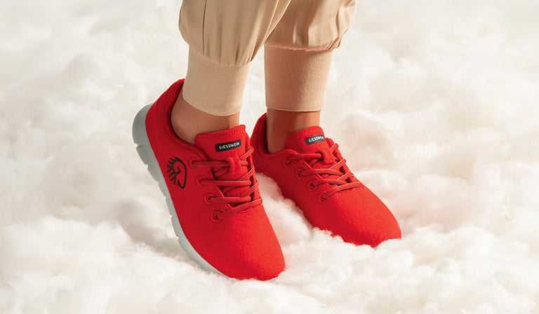 Eine Person trägt leuchtend rote Giesswein-Sneaker mit grauer Sohle (Merino Runners von Giesswein). Die Schuhe bestehen aus weichem Stoff und haben eine Schnürung. Die Person trägt beigefarbene Jogginghosen. Die Füße stehen auf einer flauschig wirkenden, weißen Unterlage, die an Wolle oder Wolken erinnert.