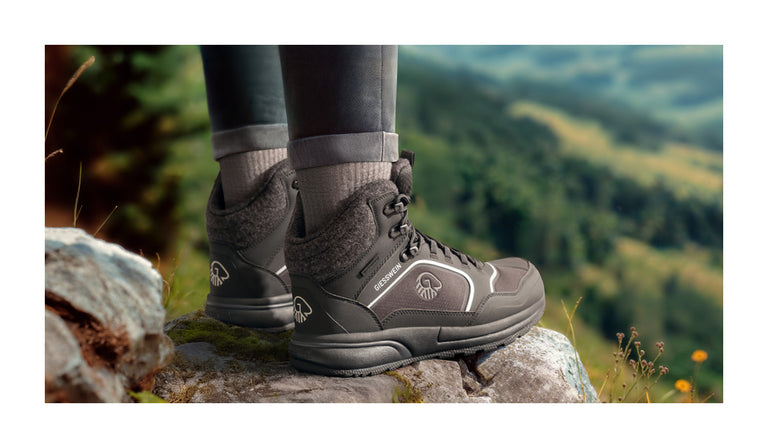 Nahaufnahme von zwei Füßen in dunkelgrauen, knöchelhohen Wanderschuhen (Wool Cross X Alpine von Giesswein) mit Wollfutter, die auf einem Felsen stehen. Die Schuhe tragen das Giesswein-Logo und befinden sich in einer grünen, leicht verschwommenen Berglandschaft mit Blumen im Vordergrund.
