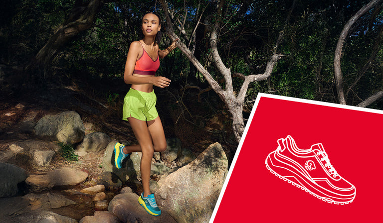 Eine sportliche Frau joggt durch einen schattigen Waldpfad mit Felsen und Baumstämmen. Sie trägt ein rotes Sporttop, neongrüne Shorts und auffällige blau-gelbe Laufschuhe (Wool Trail X von Giesswein). Im Vordergrund ist ein rotes Rechteck mit der weißen Illustration eines Sportschuhs und dem Logo „GIESSWEIN“ zu sehen.