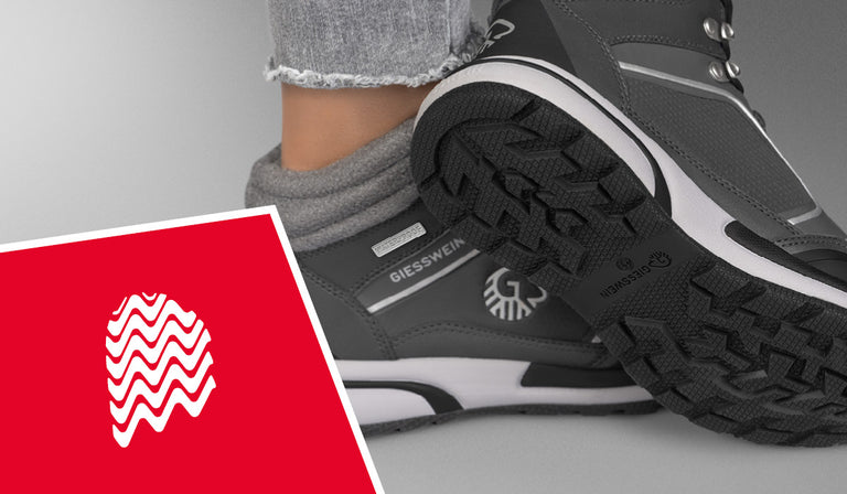 Nahaufnahme eines grauen Wanderschuhs (Wool Cross X Alpine von Giesswein) mit dicker, stark profilierter Gummisohle und sichtbarem GIESSWEIN-Logo an der Seite und auf der Sohle. Der Schuh wird von einer Person mit hochgekrempelter grauer Jeans und grauen Wollsocken getragen. Im Bild befindet sich ein rotes Quadrat mit einem stilisierten, weißen Fingerabdruck aus gewellten Linien – Symbol für Profil oder Griffigkeit.