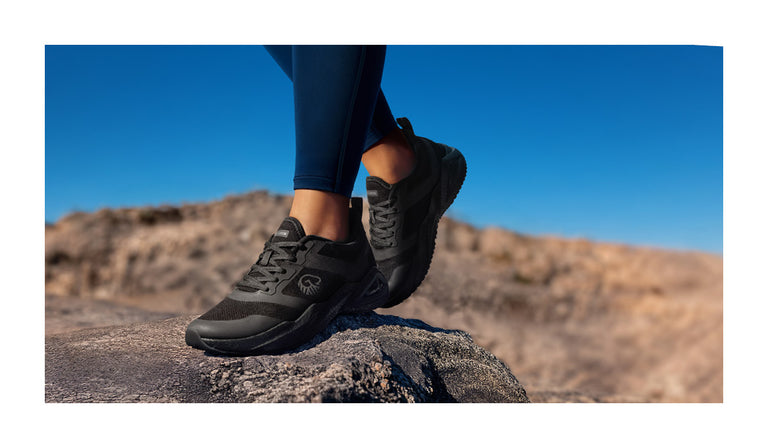 Person steht auf einem Felsen in einer felsigen, trockenen Landschaft unter klarem, blauem Himmel. Sie trägt schwarze Trail-Schuhe mit robuster Sohle (Wool Peak von Giesswein) und dunkler Sportleggings – bereit für eine Wanderung oder Outdoor-Aktivität.