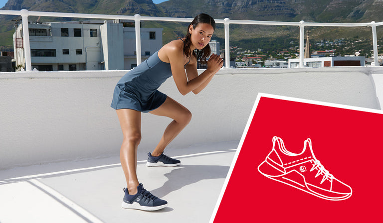 Frau in dunkelblauem Sportkleid macht eine Kniebeuge auf einer sonnigen Dachterrasse mit Blick auf eine Stadt und bewaldete Hügel. Sie trägt dunkelblaue Sportschuhe mit weißer Sohle (Wool Trainer von Giesswein). In der rechten unteren Ecke ist eine rote Fläche mit weißer Schuh-Illustration eingeblendet.