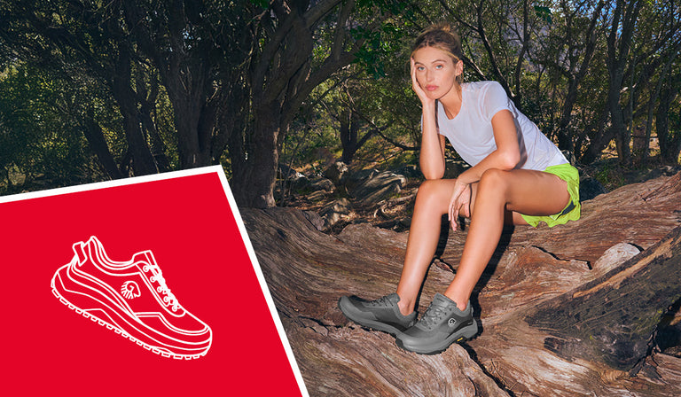 Frau sitzt entspannt auf einem großen Baumstamm im Wald und trägt ein weißes T-Shirt, eine neonfarbene Shorts und graue Wanderschuhe (Wool Trail X von Giesswein). Im Hintergrund dichter Baumbestand mit Sonnenlicht zwischen den Ästen. Links unten im Bild befindet sich eine rote Fläche mit der weißen Illustration eines Wanderschuhs.