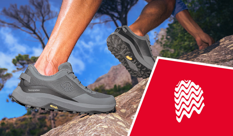 Nahaufnahme von zwei Beinen, die in grauen Wanderschuhen (Wool Trail X von Giesswein) einen steilen Felsen erklimmen. Die Schuhe haben ein robustes Profil und tragen das Logo ‚Waterproof‘ sowie eine Vibram-Sohle. Im Hintergrund blauer Himmel, Bäume und felsige Landschaft. In der rechten unteren Ecke befindet sich eine rote Fläche mit einem stilisierten weißen Schuhprofil.