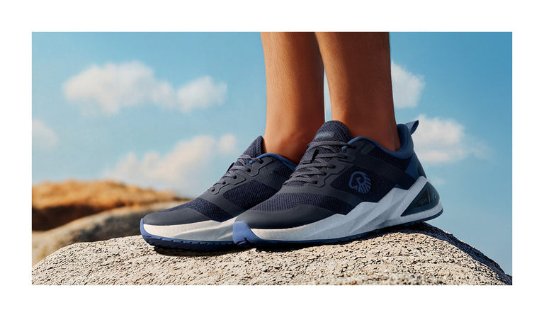 Nahaufnahme eines Paars dunkelblauer Sportschuhe (Wool Peak von Giesswein) auf einem hellen Felsen bei Tageslicht. Die Schuhe haben eine weiße, dicke Zwischensohle und ein sportliches Mesh-Design. Im Hintergrund ein klarer Himmel mit wenigen Wolken und sandfarbene Felsen in der Ferne.