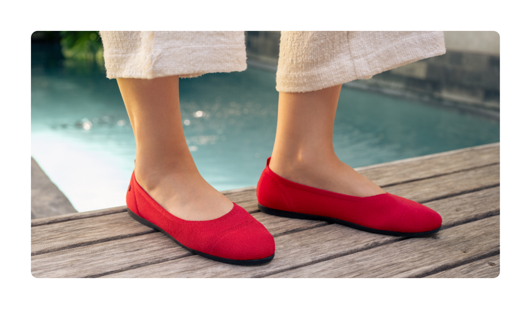 Nahaufnahme der Füße einer Person, die auf einer Holzterrasse neben einem Pool steht. Die Person trägt rote Ballerinas (Eco Ballerinas von Giesswein) und eine helle, weit geschnittene Sommerhose.