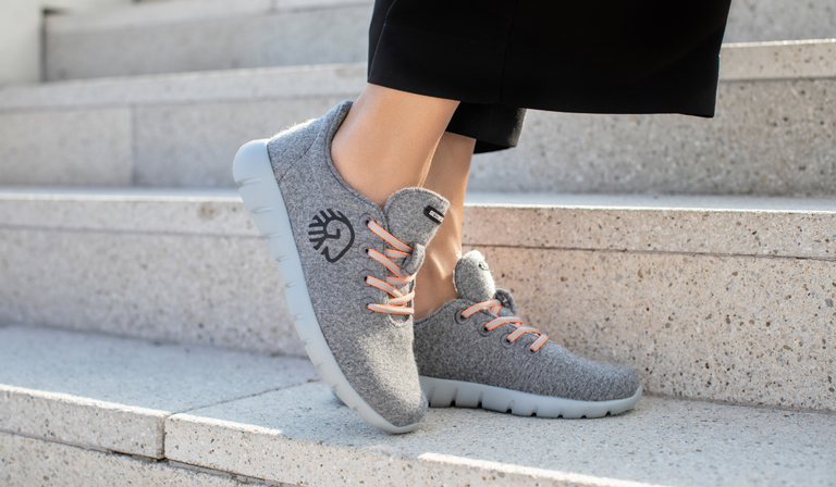 Die Füße einer Person stehen auf einer Treppenstufe. Sie trägt graue Sneakers (Merino Runners von Giesswein) mit weißen Sohlen und orangen Schnürsenkeln. Auf den Schuhen ist das Giesswein-Logo zu sehen. Die Person trägt eine schwarze Hose, und im Hintergrund sind die Stufen der Treppe sichtbar.