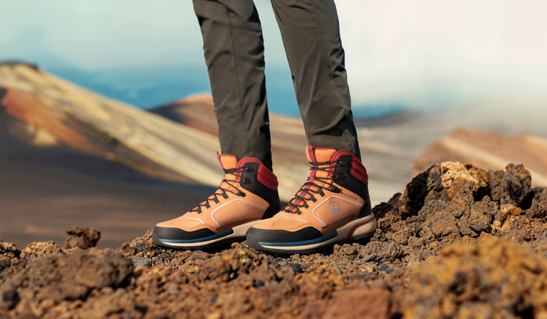 Nahaufnahme von orange-schwarzen Wanderstiefeln auf vulkanischem Gestein. Die Person trägt olivgrüne Wanderhosen und robuste Mid-Cut-Stiefel mit orange-schwarzer Farbgebung und roten Schnürsenkeln. Der Untergrund besteht aus rauem, porösem Vulkangestein in Brauntönen. Im unscharfen Hintergrund sind farbige Gesteinsformationen in warmen Erdtönen unter bewölktem Himmel zu erkennen.