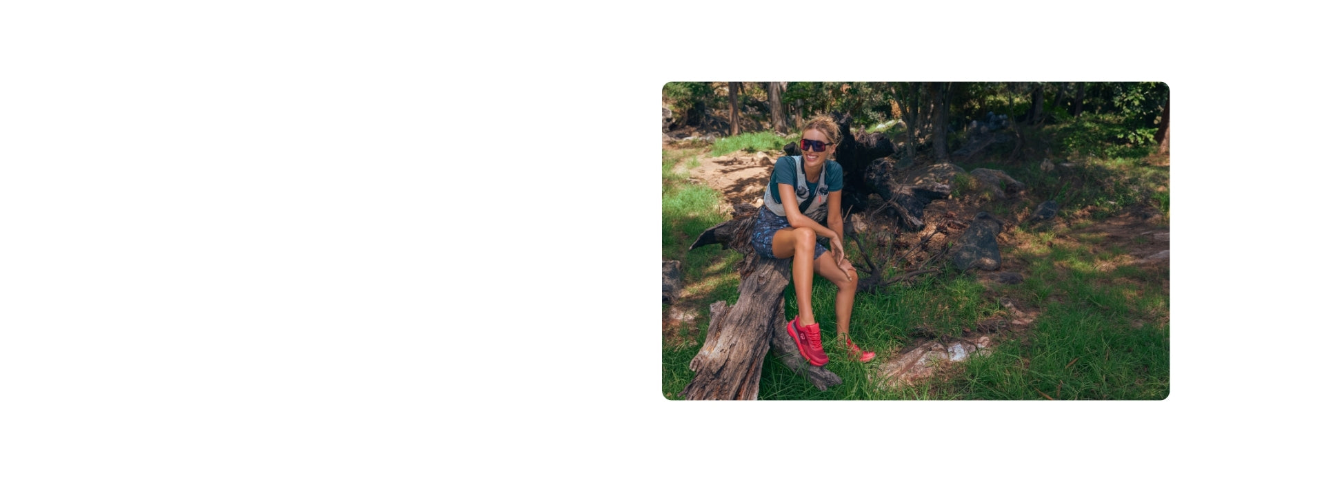Lächelnde Frau mit Sonnenbrille in grauem T-Shirt und gemusterter Shorts mit roten Sneakers sitzt auf Baumstamm in natürlicher Waldumgebung mit Steinen und Gras.