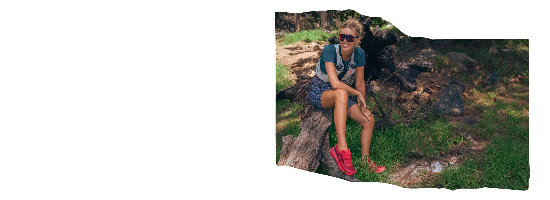 Eine lächelnde Frau sitzt entspannt auf einem Baumstumpf im Wald. Sie trägt sportliche Kleidung, eine Sonnenbrille und leuchtend rote Wanderschuhe (Wool Trail X von Giesswein). Um sie herum liegt Waldboden mit Gras, Steinen und Wurzeln. 