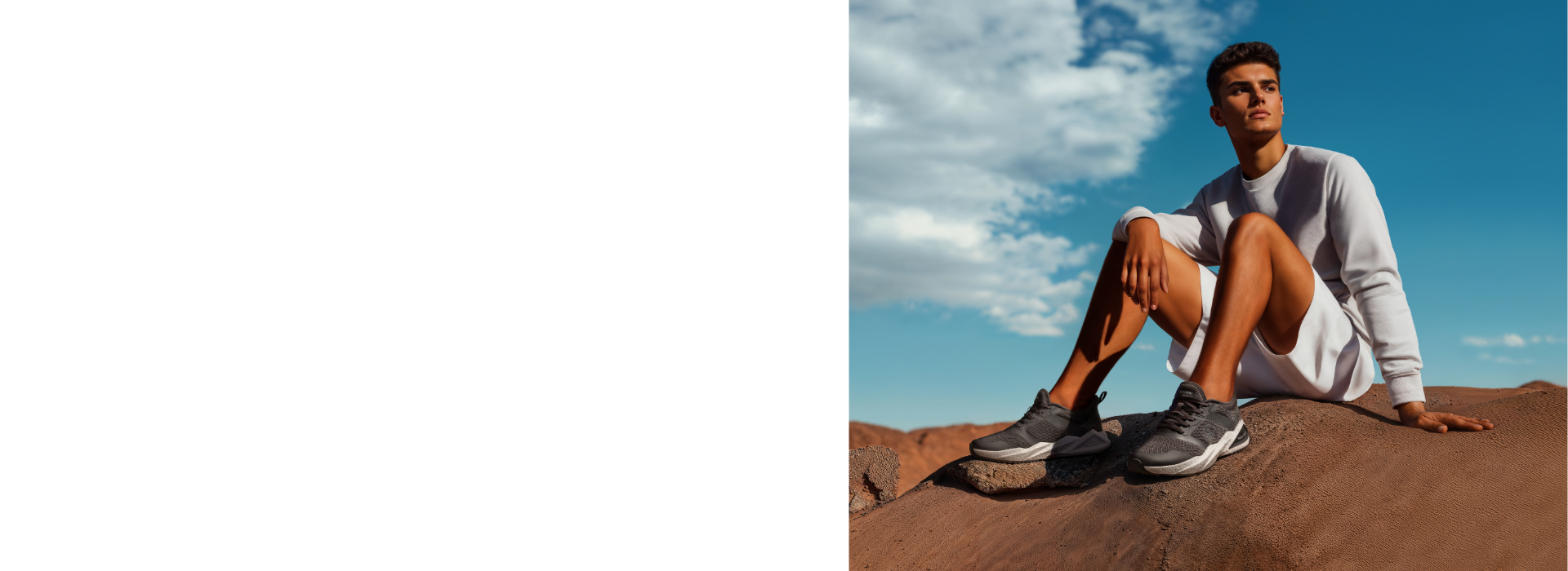 Ein junger Mann sitzt auf einem Felsen in einer Wüstenlandschaft, trägt ein weißes Sweatshirt und eine weiße Shorts. Er hat schwarze Sneakers (Wool Peak von Giesswein) an und schaut in die Ferne. Der Himmel ist blau, mit ein paar Wolken, und der Hintergrund zeigt eine trockene, bergige Landschaft.