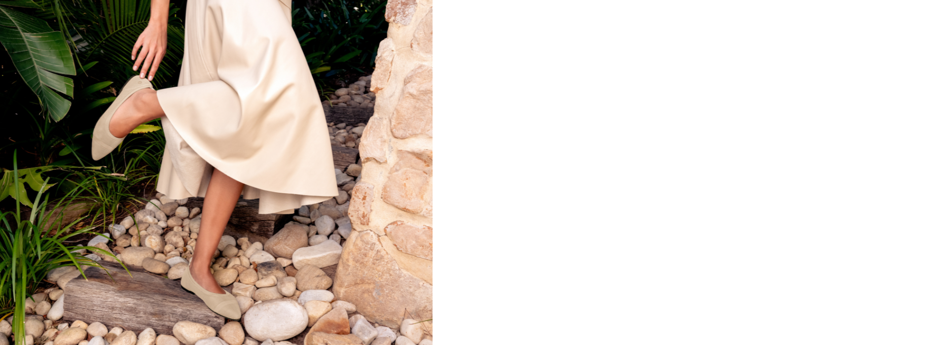 Eine Person mit cremefarbenem, fließendem Kleid steigt barfuß in beigefarbenen Ballerinas (Eco Ballerinas von Giesswein) eine kleine Steintreppe hinauf. Die Szene spielt im Freien zwischen tropischem Grün und einer Natursteinmauer und wirkt sommerlich und elegant.