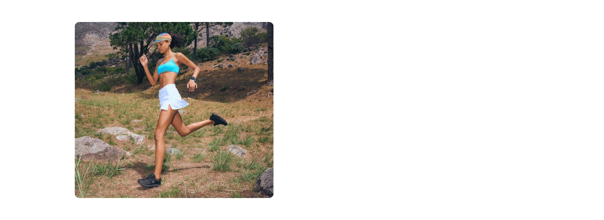 Eine sportlich gekleidete Frau joggt über eine Wiese mit Felsen und trockenem Gras. Sie trägt einen türkisfarbenen Sport-BH, einen weißen Laufrock, schwarze Trailrunning-Schuhe (Wool Trail X von Giesswein) und eine bunte Sonnenblende. Im Hintergrund sind Bäume, Büsche und ein Hang mit Felsen zu sehen.