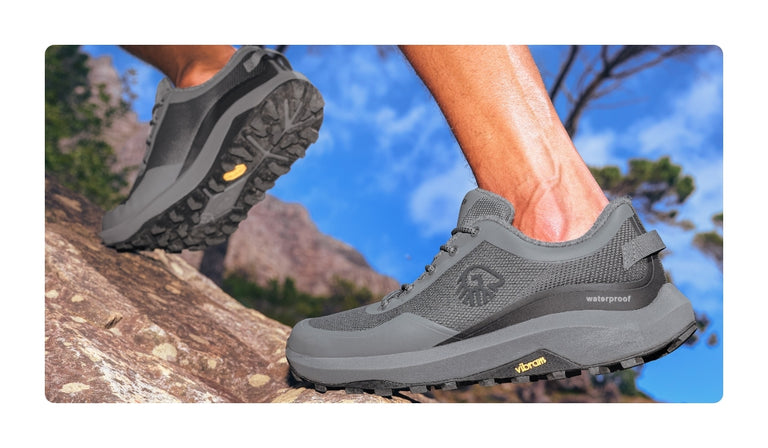 Nahaufnahme eines grauen Wanderschuhs (Wool Trail X von Giesswein) mit tiefem Profil auf einem steinigen Hang. Die Schuhe haben eine Vibram-Sohle und sind als „waterproof“ gekennzeichnet. Ein Bein ist beim Auftritt zu sehen, im Hintergrund zeigen sich blauer Himmel, Felsen und Bäume.