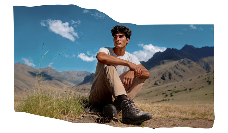 Ein junger Mann sitzt in beiger Wanderkleidung und grauen Trekkingstiefeln (Wool Cross X Alpine von Giesswein) im Gras einer weiten Berglandschaft. Er blickt nachdenklich in die Ferne. Der Himmel ist blau mit vereinzelten weißen Wolken, im Hintergrund erheben sich karge, felsige Berghänge.