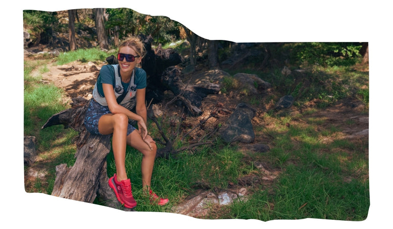 Eine lächelnde Frau sitzt entspannt auf einem Baumstumpf im Wald. Sie trägt sportliche Kleidung, eine Sonnenbrille und leuchtend rote Wanderschuhe (Wool Trail X von Giesswein). Um sie herum liegt Waldboden mit Gras, Steinen und Wurzeln. 