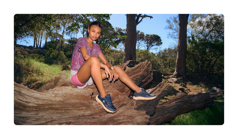 Eine junge Frau sitzt entspannt auf einem großen umgestürzten Baumstamm im Wald. Sie trägt ein sportliches Outfit mit pink-violettem Shirt, rosa Shorts und dunkelblauen Outdoor-Sneakern mit heller Sohle (Wool Trail X von Giesswein). Im Hintergrund sind Bäume, Felsen und sonnige Waldlandschaft zu sehen. 